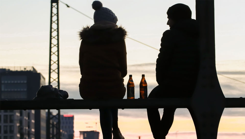 The silhouettes of a woman and a man against the sunset, with two bottles of beer between them (Photo: birgit on unsplash)