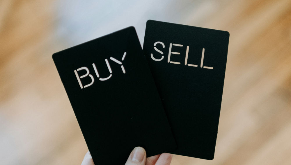 Hand holding two black cards saying “buy” and “sell” (Photo by Kelly Sikkema on Unsplash)
