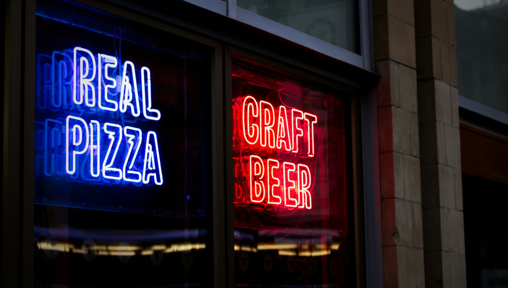 Two neon signs next to each other, saying “Real Pizza” and “Craft Beer” (Photo: Leigh Cooper on Unsplash)