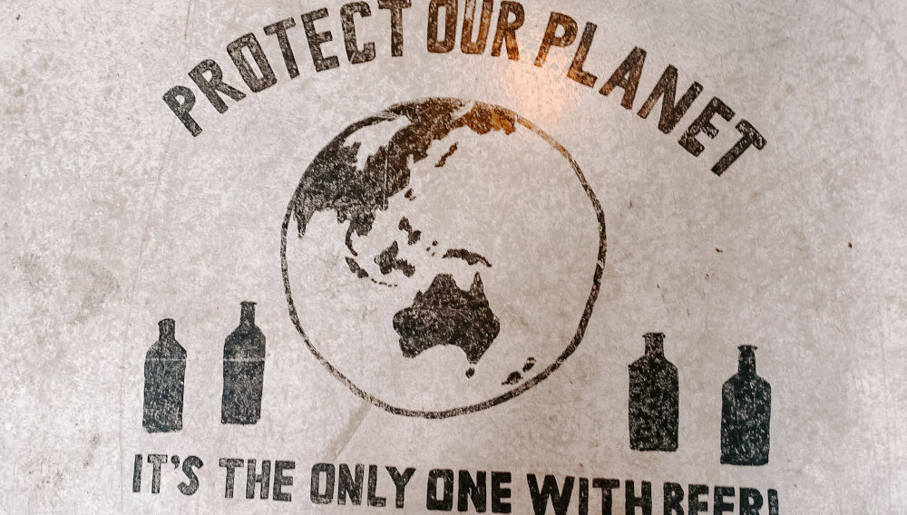 Sign on concrete floor, saying: Protect our planet, it’s the only one with beer (Photo by S O C I A L . C U T on Unsplash)