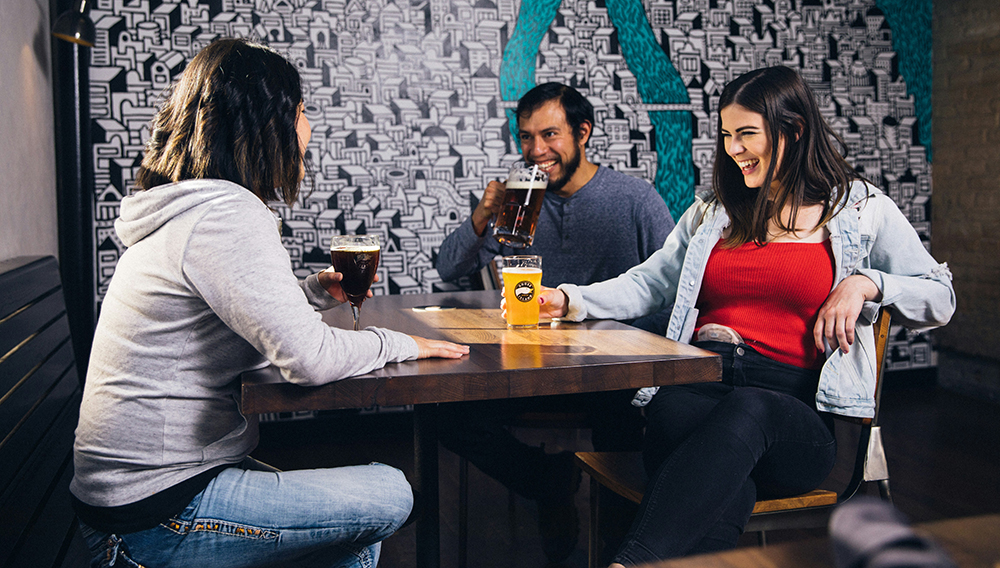 Drei Leute, die an einem Tisch sitzen, Bier trinken und lachen (Foto: Elevate auf Unsplash)